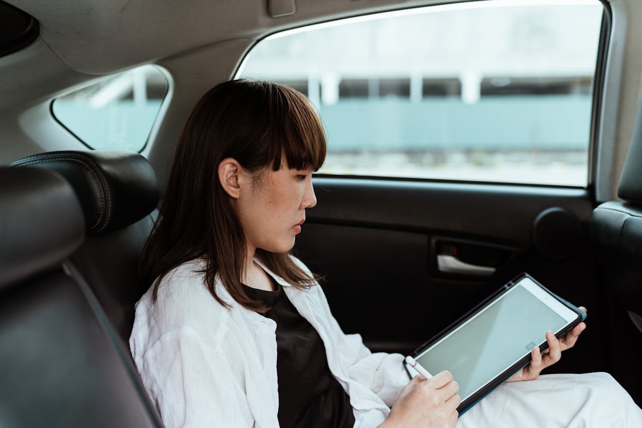 Nosotros Side view serious Asian female in casual outfit using tablet with empty screen and stylus while riding in car backseat on daytime